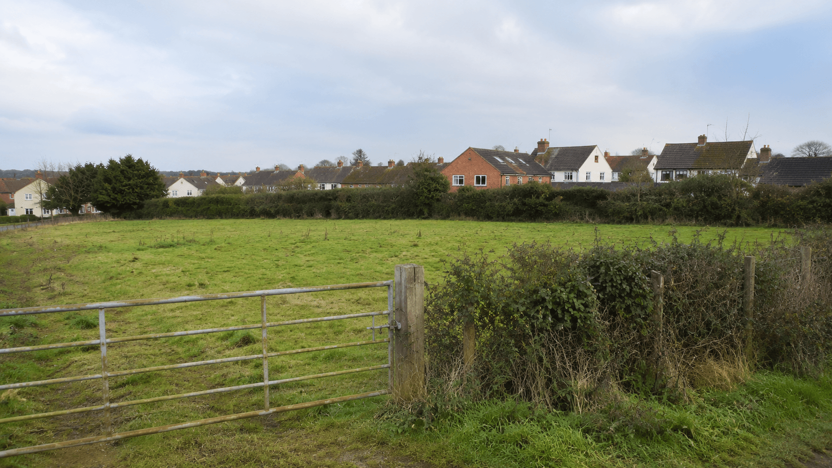 A small grass paddock with a wooden gate and hedgerow, with semi-detached houses just beyond the boundary.