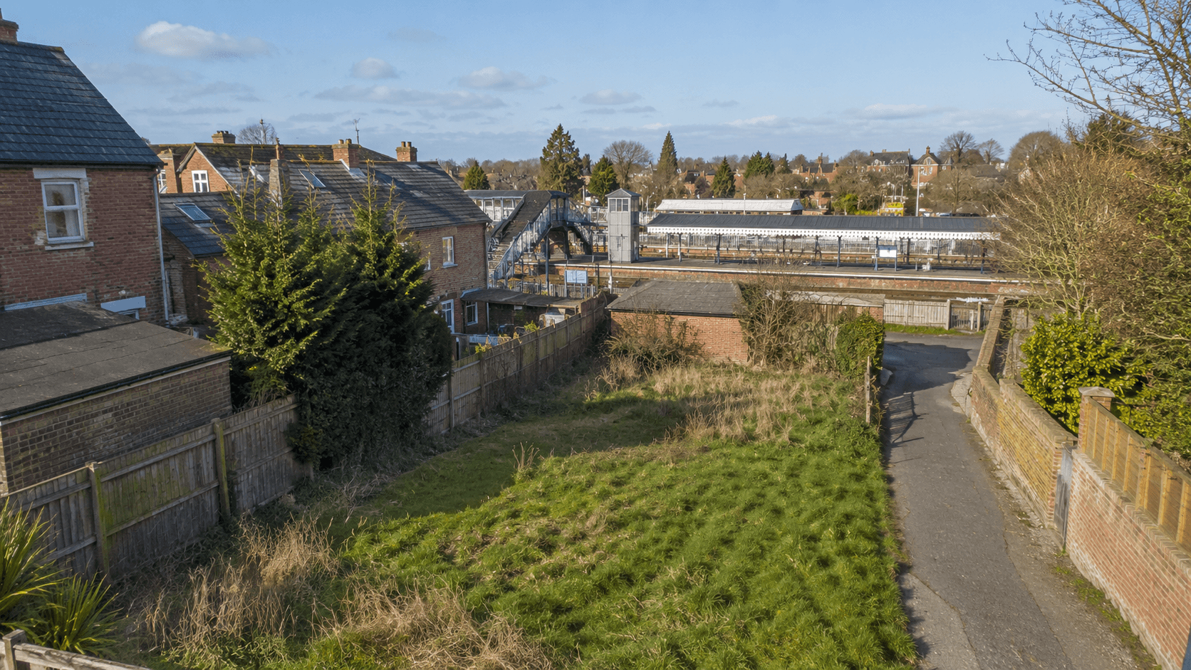 A walled grass plot looking towards a railway station with platform canopies and trains visible beyond the fence.