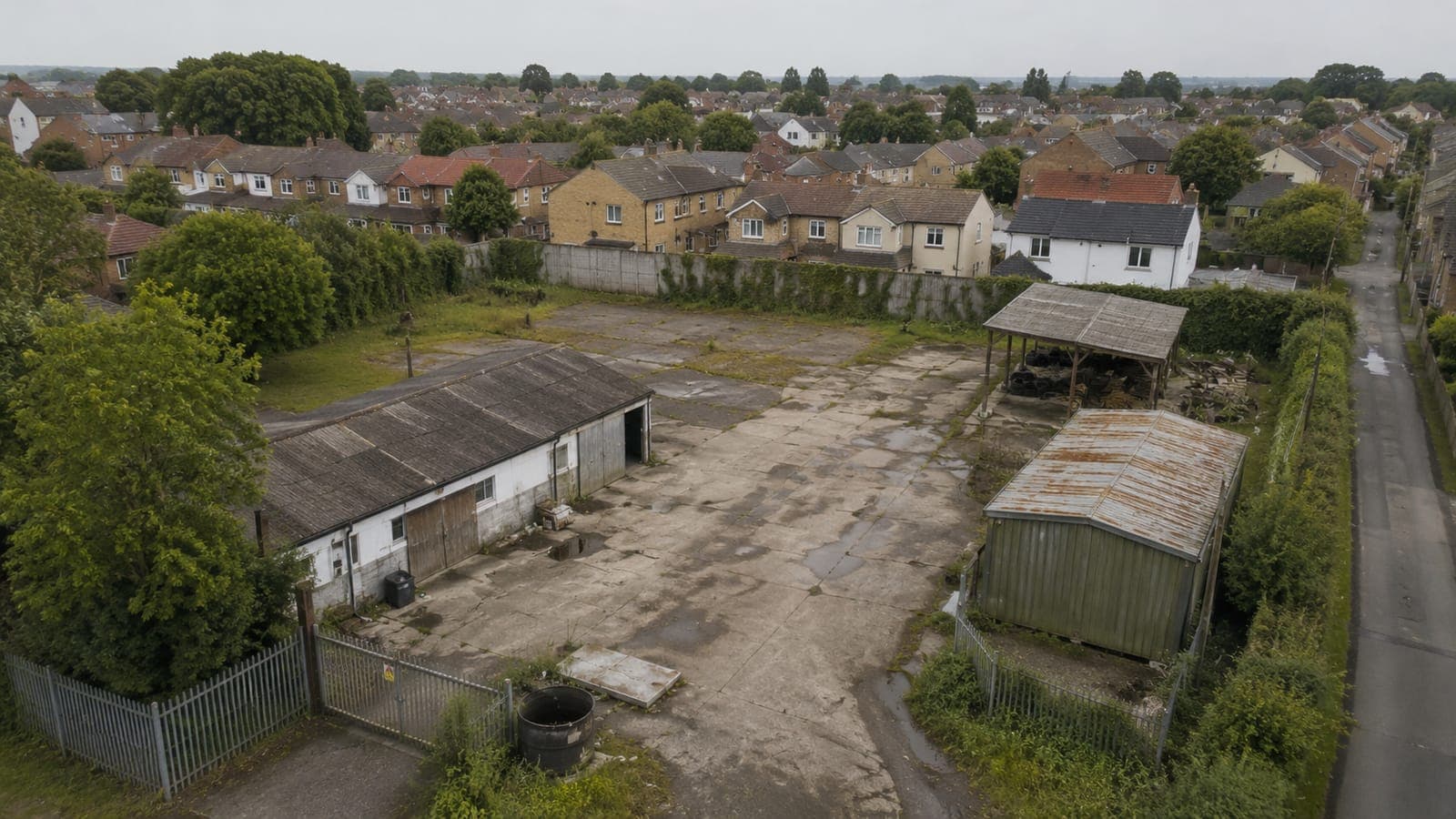 An aerial view of a derelict yard with two outbuildings and palisade fencing, with residential rooftops behind.